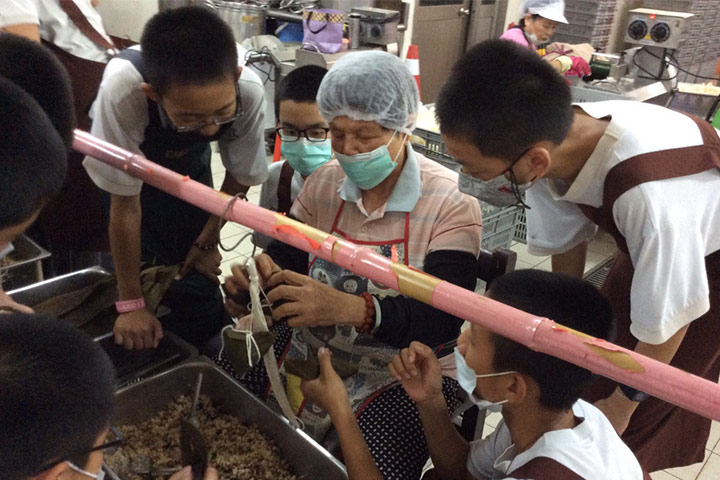 福智教育園區的廚房內，阿嬤義工正努力傳遞包粽子的手藝和用心，學生們在一旁專注地學習。